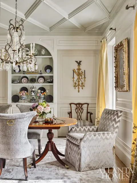 Dining room with a crystal chandelier, coffered ceiling, built-in shelves displaying decorative plates, and a round wooden table with upholstered chairs.