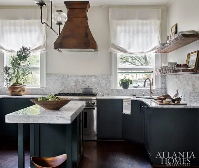 Dark green kitchen cabinets with white marble countertops and backsplash, a copper range hood, and two windows with white roman shades.