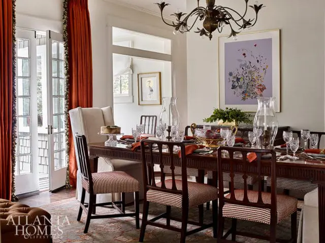 Formal dining room with a dark wood table set for a meal, upholstered chairs with striped fabric, tall windows with orange drapes, and a large floral artwork on the wall.