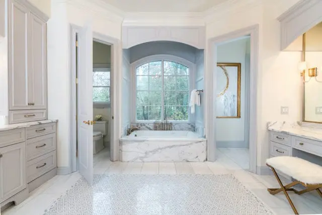 Light gray bathroom with marble bathtub, white cabinetry, and a vanity with a fur-covered stool. A doorway reveals a toilet.