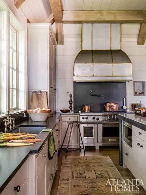Farmhouse kitchen with white shaker cabinets, dark countertops, stainless steel range with custom hood, and exposed wooden beams.