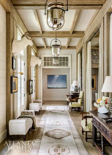Hallway with shiplap walls, exposed wood beams, and large pendant lights. Taxidermied goat heads mounted on the wall, with upholstered benches and a patterned runner on the wood floor.
