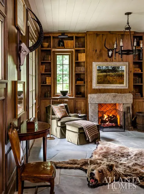 Library with wood paneled walls, stone fireplace with a roaring fire, plush green armchair with ottoman, and a bear skin rug on the floor.