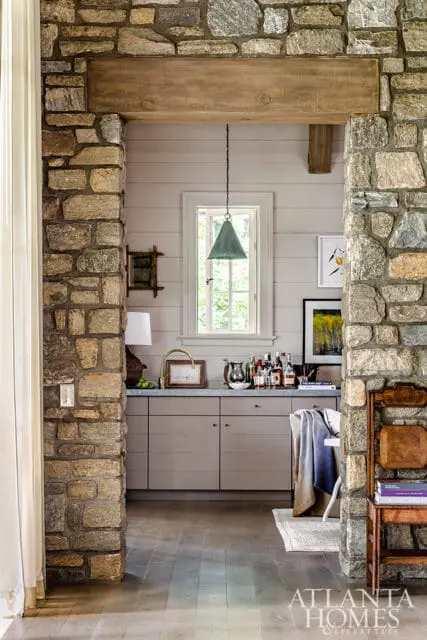 Stone pillars frame a bar area with grey cabinets, a marble countertop, and a window above. A green pendant light hangs overhead.