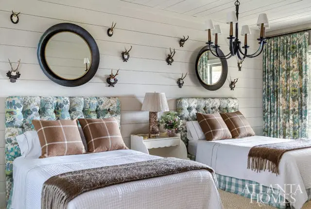 Bedroom with two queen beds featuring tufted headboards with a floral and animal print fabric. The walls are paneled in white shiplap and decorated with mounted antlers and two round mirrors.