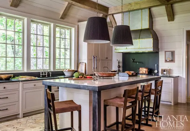 Farmhouse kitchen with white paneled walls, exposed wooden beams, a large island with a metal frame and marble countertop, and dark pleated pendant lights.