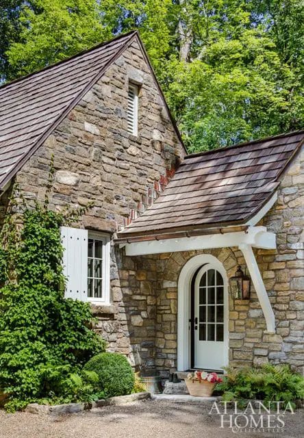 Stone cottage with a gabled roof, arched white door with a glass pane, and white shutters on a window. Lush green foliage surrounds the entrance.