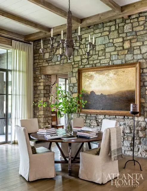 Dining room with a round wooden table, upholstered chairs, stone accent wall, large landscape painting, and a wrought iron chandelier.