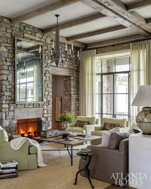 Living room with stone fireplace, exposed wood beams, and light green upholstered furniture. A large mirror hangs above the fireplace.