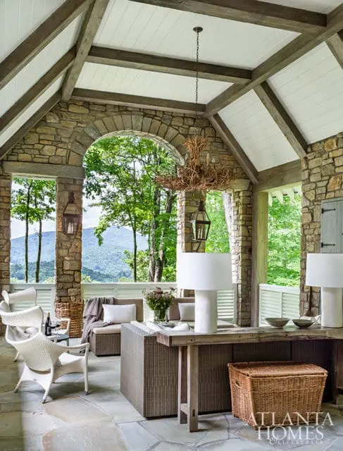 Covered porch with stone walls, wood-beamed ceiling, and large windows overlooking mountains. Furnished with wicker chairs, a sectional sofa, and a rustic console table with lamps.