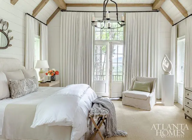 Bedroom with white shiplap walls, exposed wooden beams, and floor-to-ceiling white curtains. A large bed with white linens is center, with a small armchair and a sculpture on a pedestal to the right.