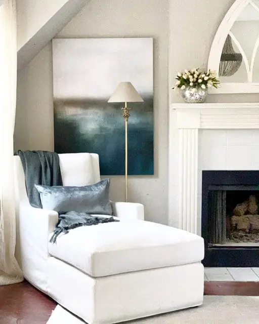 White chaise lounge with blue velvet pillow and gray throw blanket next to a fireplace and abstract art.
