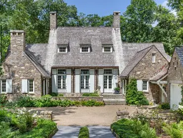 Stone cottage with a grey shake roof, multiple dormer windows, and light blue shutters. Lush green landscaping surrounds the home.