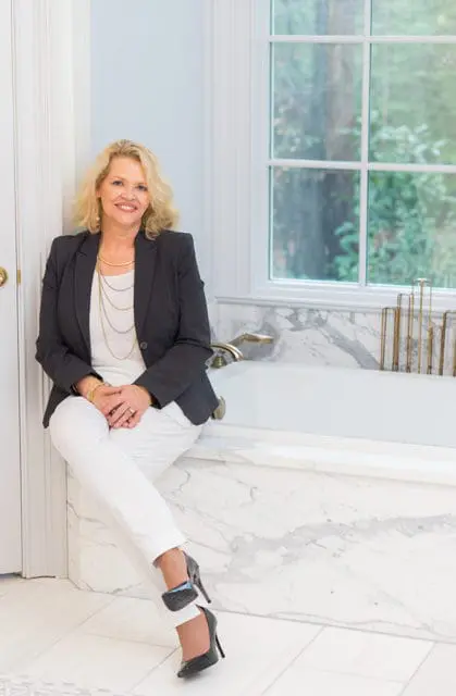 Woman with blonde hair wearing a dark blazer, white top, and white pants, sitting on the edge of a marble bathtub.