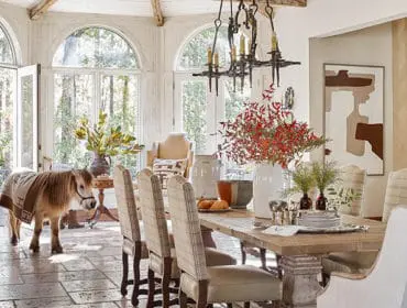 Sunroom with stone tile floor, large arched windows overlooking trees, a rustic wooden dining table set with plaid chairs, and a miniature horse wearing a blanket