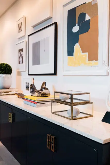 Dark blue cabinetry with brass hardware topped with a marble countertop displaying stacked glass boxes with stones, books, and artwork on the wall above.