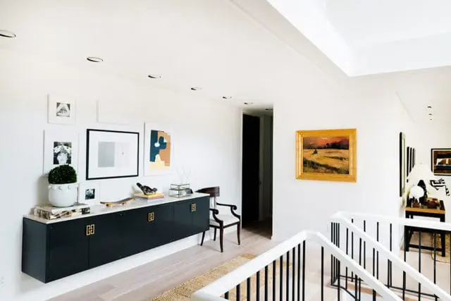 Hallway with white walls, a black floating credenza with gold hardware, and a gallery wall of framed art. A landscape painting hangs on the adjacent wall.