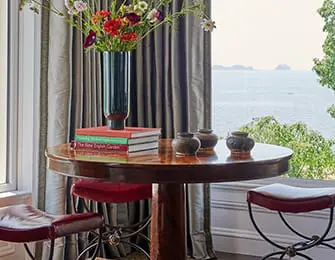Round wooden table with stacked books and vases, flanked by two red stools, with a window view of the ocean and islands.