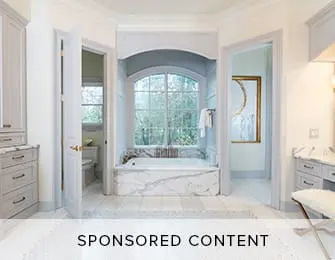 Spacious bathroom with a freestanding marble bathtub under a large arched window, flanked by grey cabinetry and doors leading to other rooms.