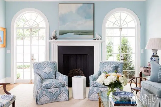 Light blue living room with a black fireplace, two upholstered armchairs with blue and white damask pattern, and arched French doors with white mullions.