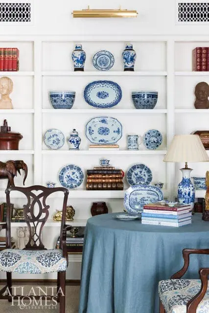 White built-in shelving displaying blue and white porcelain plates, vases, and bowls, with decorative busts and books. A round table draped in a blue cloth sits in the foreground, next to a chair with patterned upholstery.
