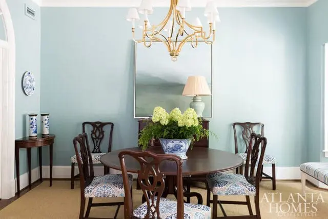Light blue dining room with a dark wood round table, upholstered chairs with blue and white patterned fabric, a gold chandelier, and a landscape painting above a dresser.
