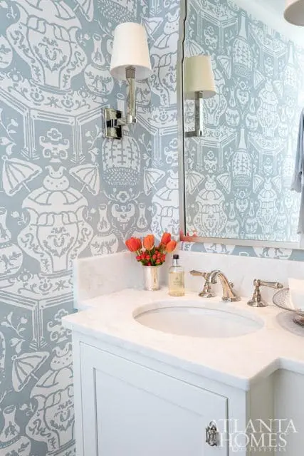 Powder room with blue and white Chinoiserie wallpaper, white marble vanity countertop, and polished nickel faucet. A silver vase holds orange tulips.