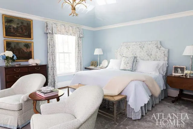 Light blue bedroom with a floral upholstered headboard, white bedding, two upholstered armchairs, a wooden dresser with paintings, and a gold chandelier.
