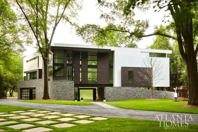 Modern multi-story home with dark wood siding, white stucco, and stone accents. Features large windows, a glass garage door, and a driveway with stone pavers.