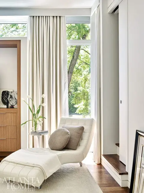 Cream chaise lounge with two neutral throw pillows and a plaid throw blanket, next to a small side table with a vase of white tulips. Large windows with cream curtains overlook a tree-filled yard.