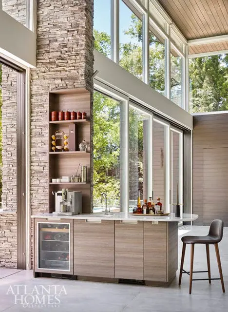 Modern bar area with a stone accent wall, wood cabinetry, marble countertop, wine fridge, espresso machine, and a tray of liquor bottles. Large windows offer views of lush greenery.