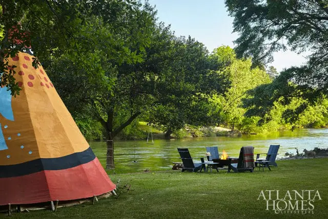 Teepee and Adirondack chairs with a fire pit by a river, surrounded by lush green trees.