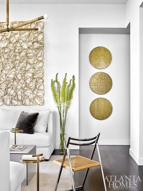 Modern living room with white sofa, abstract woven wall art, and gold circular wall decor. A chrome and cane chair sits on dark wood floors.