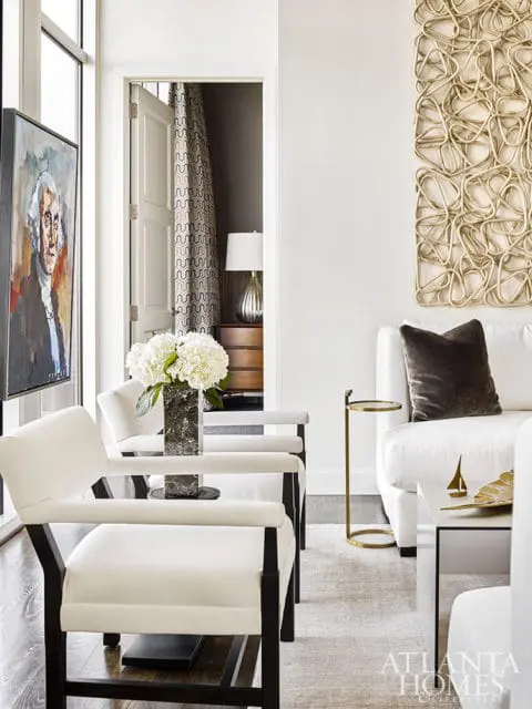 Modern living room with white armchairs, a white sofa, and a large abstract wall sculpture. A portrait hangs on the left wall.
