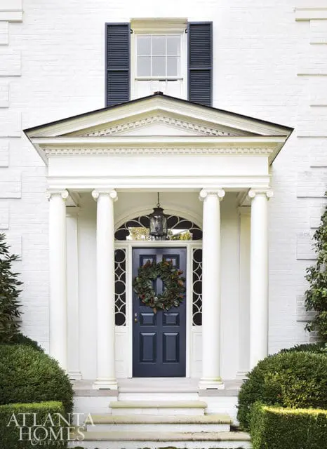 White brick house with a dark blue front door, a magnolia wreath, and a pediment supported by columns.