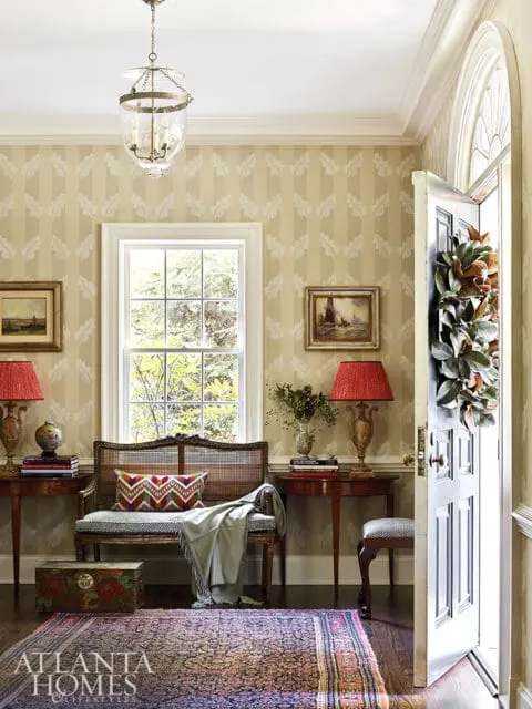 Entryway with striped wallpaper, a cane bench with patterned pillows and a throw, a vintage trunk, and a Persian rug. A door with a magnolia wreath is open to the right.