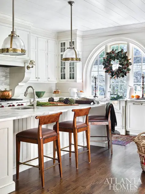 White kitchen with marble countertops, wood bar stools with leather seats, and a large arched window adorned with a magnolia wreath.