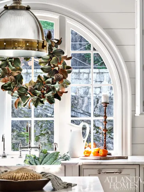 White kitchen with a large arched window, a magnolia wreath, a farmhouse sink with kale, a white pitcher, and a wooden cutting board with bell peppers.