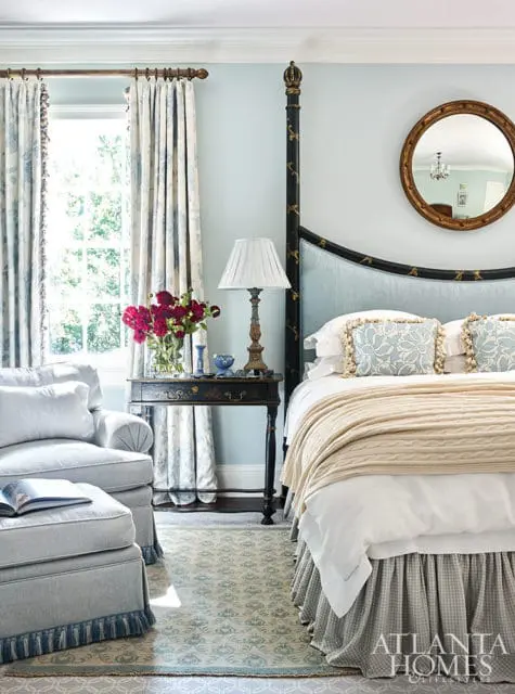 Light blue bedroom with a black and gold four-poster bed, upholstered headboard, white bedding, and a cream cable-knit throw. A patterned rug, armchair with ottoman, and a bedside table with a lamp and flowers are visible.