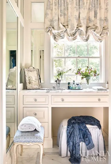Cream vanity with marble countertop, drawers, and a makeup area. A window with a floral balloon shade is above. Two stools, one with a folded towel and the other draped with a blue scarf, are in front.
