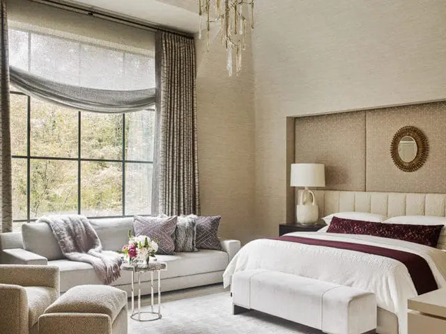 Bedroom with a large window overlooking trees, a king-sized bed with white linens and a burgundy accent runner, and a grey sofa with plush pillows and a throw blanket.