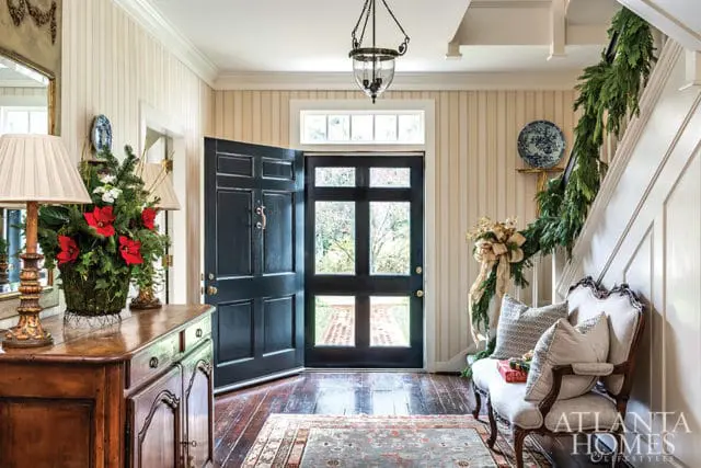 Entryway with striped wallpaper, a dark blue paneled door, a wooden console table with floral arrangements, a bench with decorative pillows, and a staircase adorned with garland.
