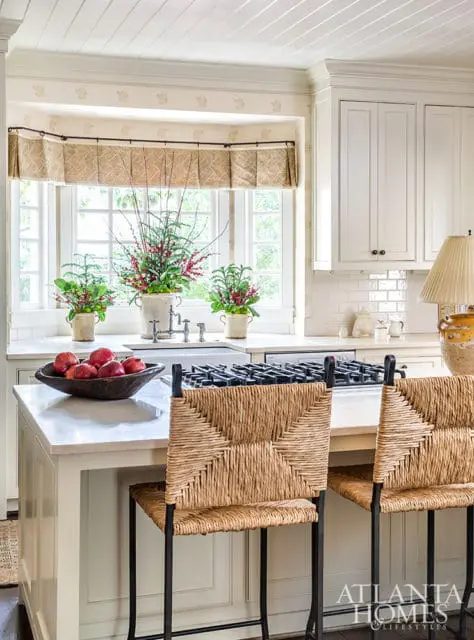 Cream kitchen with white subway tile backsplash, bay window with floral valance, and woven bar stools at marble island with bowl of apples.