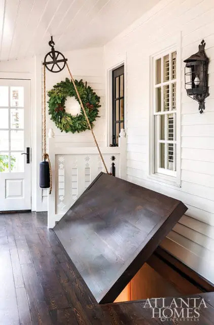 Dark wood floor with an open trapdoor revealing a lit space below, a rope and pulley system hangs above a festive green wreath on a white paneled wall.