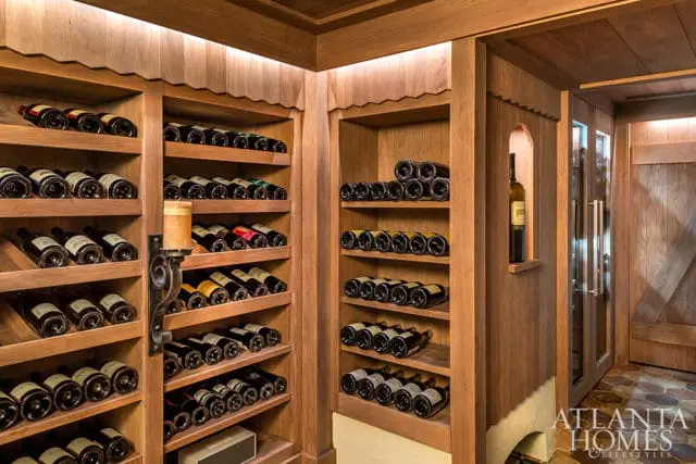 Wooden wine cellar with floor-to-ceiling shelves filled with wine bottles, built-in display niche, and hexagonal tile flooring. Atlanta Homes & Lifestyles logo in the lower right corner.