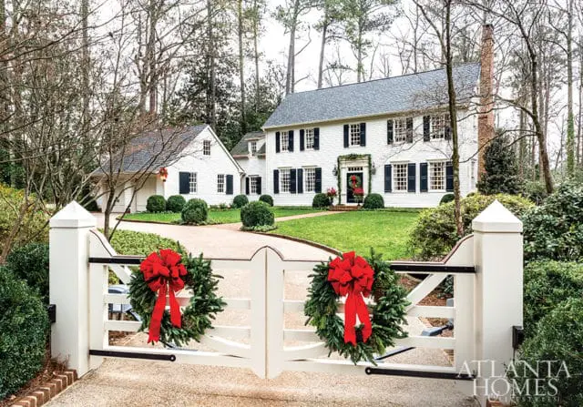 White colonial-style house with black shutters and a brick chimney, decorated for Christmas with wreaths and garland. A white gate with two wreaths and red bows guards the driveway.