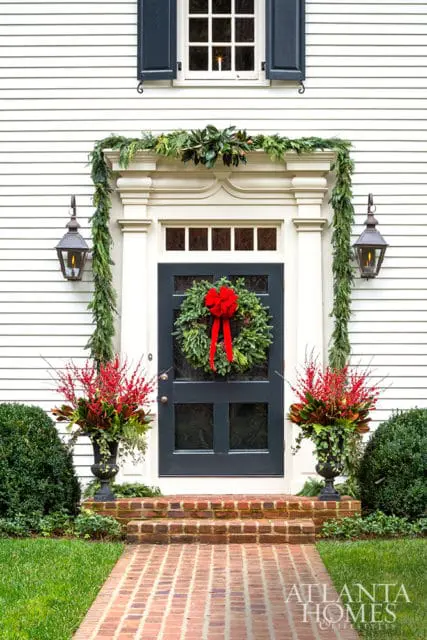 Dark gray front door decorated with a lush green wreath and red bow, flanked by urns filled with red berries and greenery. A garland of evergreen boughs drapes over the doorway, leading to a brick pathway and manicured lawn.