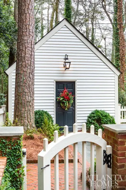 White clapboard shed with a dark gray door decorated with a festive wreath, flanked by manicured bushes and a large pine tree. A white picket fence and arched gate lead to a brick pathway.