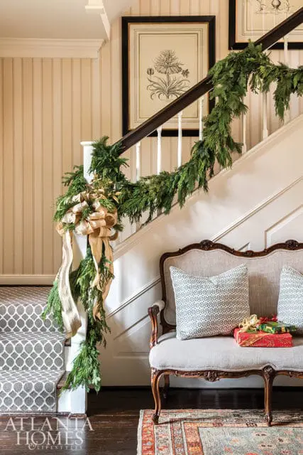 Foyer with staircase decorated for Christmas, featuring green garland, a large gold bow, framed botanical prints, a settee with patterned pillows, and wrapped gifts.