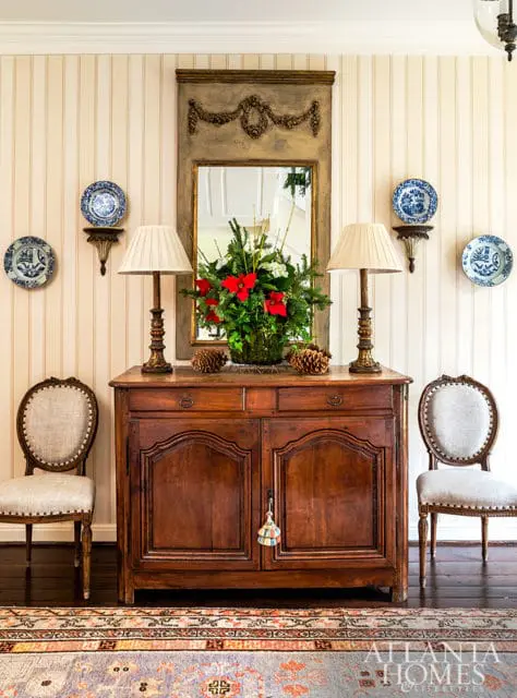 Antique wooden sideboard with two lamps, a floral arrangement, and decorative plates on striped wallpaper. Two chairs flank the sideboard.
